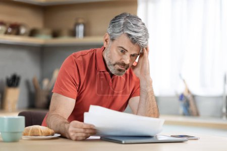 Photo for Crisis 2022. Unhappy middle aged male entrepreneur reading correspondence while having breakfast at home, sad man sitting at kitchen desk with laptop and smartphone on, holding documents, copy space - Royalty Free Image