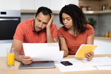 Photo for Despaired millennial black family in red t-shirts work with documents, check bills, have problems with calculate bookkeeping in kitchen interior. Stress, crisis at home, debts, credit and mortgage - Royalty Free Image