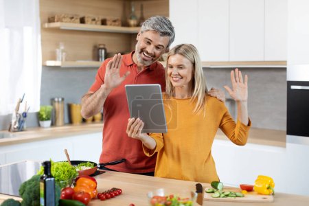 Téléchargez les photos : Joyeux conjoint d'âge moyen cuisiner dîner ensemble à la maison, avoir un appel vidéo avec la famille ou des amis, tenant tablette numérique moderne et agitant à l'écran webcam, intérieur de la cuisine confortable, espace de copie - en image libre de droit