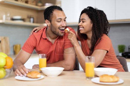 Photo for Happy millennial black wife feeds husband, couple have fun and enjoy breakfast from healthy food at weekend in kitchen interior, copy space. Love, family relationship, good morning at home together - Royalty Free Image