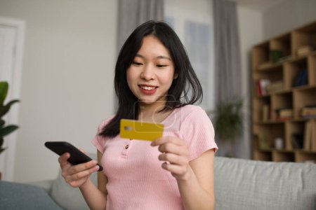 Photo for Young korean woman holding credit card and smartphone, enjoy online shopping in living room interior, free space. New app, banking, finance and cashback from buying, huge sale at home - Royalty Free Image