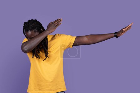 Photo for Stylish long-haired young african american man in yellow t-shirt showing cool moves dab dance pose over purple studio background, raising hands up and hiding his face, copy space - Royalty Free Image