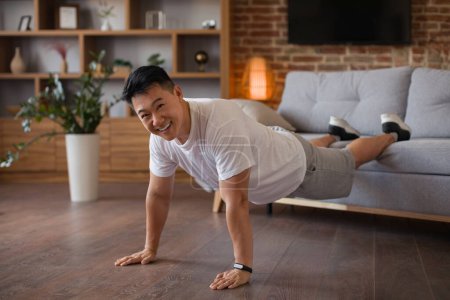 Téléchargez les photos : Fort homme asiatique d'âge moyen faisant des pompes avec les jambes sur le canapé, travaillant à la maison et souriant à la caméra. Homme exerçant les mains et les muscles abdominaux. Concept de formation domestique - en image libre de droit