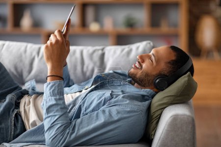 Photo for Smiling Black Man Wearing Wireless Headphones Resting With Digital Tablet At Home, Handsome Young African American Male Relaxing With Modern Gadget On Couch In Living Room, Side View - Royalty Free Image