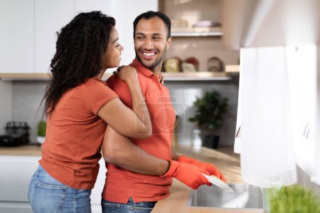 Photo for Cleaning at free time at home. Happy pretty young black woman hugging man in rubber gloves, male washes dishes in modern kitchen interior. Love, romance relationship and household chores together - Royalty Free Image