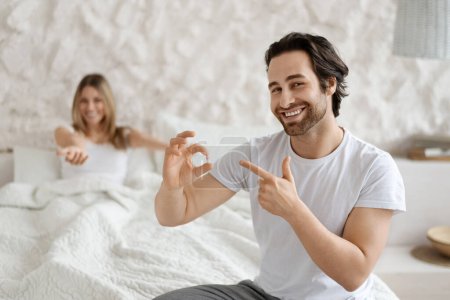 Mens health and sexual problems, male power and libido. Happy man holding pill and pointing on it, blurred smiling wife sitting in bed, close up, free space