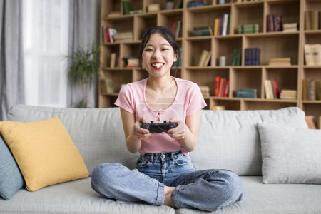 Foto de Señora coreana feliz con joystick, sentado en el sofá y jugando juegos en línea en el interior de la sala de estar, espacio de copia. Entretenimiento con dispositivo en tiempo libre en casa solo y emociones - Imagen libre de derechos