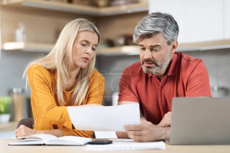 Concerned middle aged spouses grey-haired man and blonde woman sitting at desk in front of laptop, checking bills, making countings, working on family budget together, kitchen interior