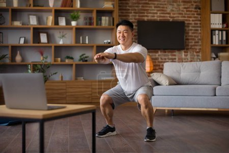 Téléchargez les photos : Coréen athlétique homme mature qui fait des squats profonds pendant l'entraînement en ligne à la maison, en regardant l'écran d'ordinateur portable. Sporty asiatique mâle exercice en face de l'ordinateur, copier l'espace - en image libre de droit