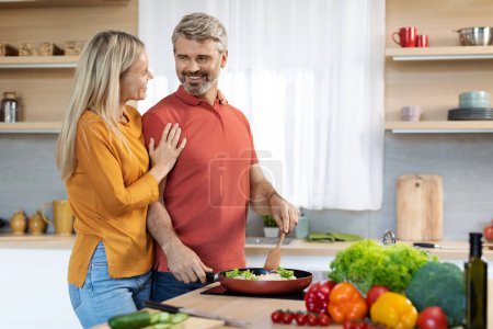 Photo for Loving beautiful blonde wife middle aged woman in casual hugging her smiling handsome husband at cozy kitchen, mature man cooking healthy delicious dinner for his family at home, copy space - Royalty Free Image