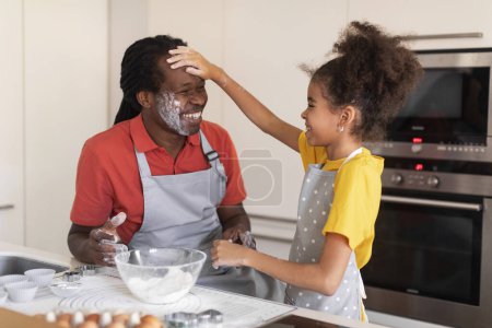 Téléchargez les photos : Joyeux père afro-américain et sa fille s'amusent pendant la cuisson dans la cuisine, mignonne Preteen Black Girl Toucher ludique Pères visage avec farine et rire, profiter de passer du temps ensemble - en image libre de droit
