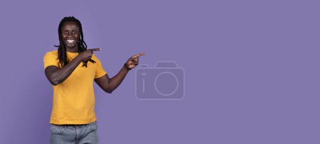 Foto de Joven afroamericano alegre fresco con las trenzas largas en la camiseta amarilla que señala el espacio de la copia para el anuncio y sonriendo sobre fondo púrpura del estudio, la tela-bandera. Bonita oferta, descuento, noticias - Imagen libre de derechos