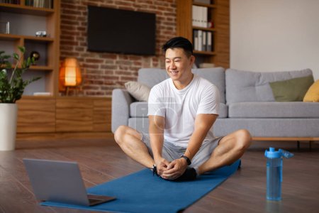 Téléchargez les photos : Pratique du yoga en ligne. mature asiatique homme faire papillon asana sur tapis de sport, suivant tutoriel vidéo sur ordinateur portable pc, copier l'espace. Coréen positif mâle exercice à la maison - en image libre de droit