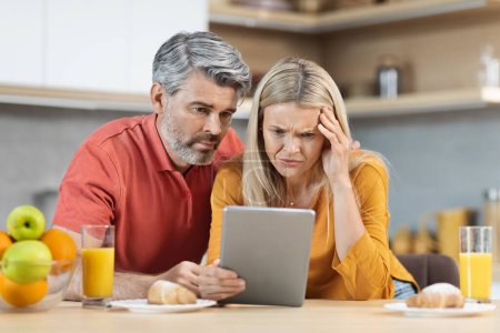 Photo for Anxious beautiful middle aged couple breakfast together at home, sitting at kitchen table, using digital tablet, looking at gadget screen with stressed face expression, reading bad news, copy space - Royalty Free Image