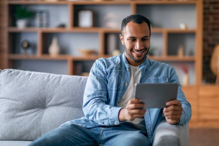 Téléchargez les photos : Joyeux mâle noir regarder des vidéos sur tablette numérique tout en se relaxant sur le canapé à la maison, souriant homme afro-américain reposant avec gadget moderne dans le salon intérieur, profiter du passe-temps domestique - en image libre de droit