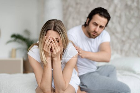 Relationship of family, problems in couple, quarrel concept. Loving caucasian man calming his crying wife, sitting on bed in light modern bedroom interior