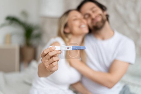 Glückliche kaukasische Ehepartner mit positivem Schwangerschaftstest und Umarmung zu Hause, selektiver Fokus. Liebevolle Eheleute, die aufgeregt sind, zukünftige Eltern zu werden. Familienplanungskonzept