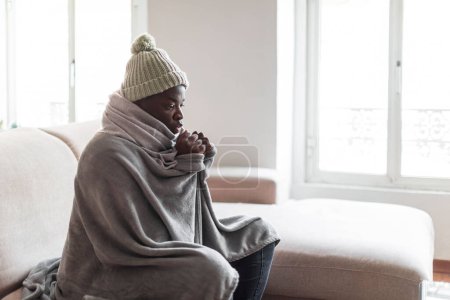Téléchargez les photos : Malheureux homme afro-américain stressé en chapeau et couverture assis dans le salon froid souffrent d'un manque de chaleur. Un jeune homme malsain lutte contre le froid glacial à la maison. Pas de concept de chauffage, espace de copie - en image libre de droit