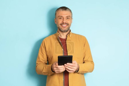 Photo for People and technology concept. Portrait of mature caucasian man holding and using tablet standing over blue studio background. Positive cheerful male adult posing with pc looking at camera - Royalty Free Image