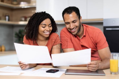 Photo for Cheerful millennial black lady and guy in red t-shirts work with documents, check bills, calculate profit in kitchen interior. Mortgages, savings and home bookkeeping at home, good news, ad and offer - Royalty Free Image