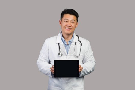 Téléchargez les photos : App pour les soins de santé à distance. Masculin coréen souriant thérapeute chinois en manteau blanc avec stéthoscope montre tablette avec écran blanc, isolé sur fond gris, studio. Conseils médicaux, services médicaux - en image libre de droit