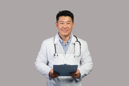 Téléchargez les photos : Smiling homme chinois mature thérapeute en manteau blanc avec stéthoscope utiliser comprimé, isolé sur fond gris, studio. Recette de médecin pour le traitement, l'examen médical, les soins de santé et le soutien en clinique - en image libre de droit