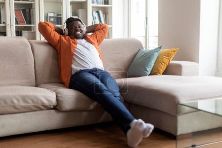 Photo for Happy cheerful chubby young black man with braids in casual enjoying music at home, sitting on couch in living room, using wireless stereo headphones, looking at copy space and smiling, full length - Royalty Free Image