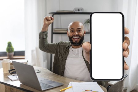 Photo for Excited latin man holding big blank smartphone and celebrating success, sitting at workplace with laptop pc, mockup. Happy male showing phone with white screen, copy space - Royalty Free Image