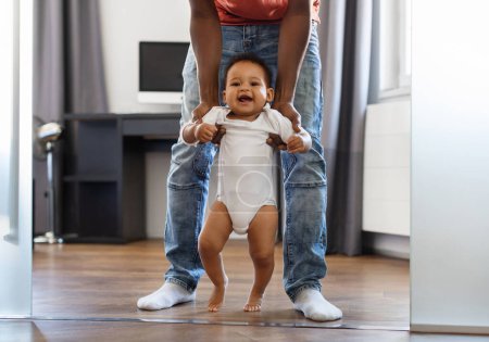 Photo for Cute black infant boy making first steps at home with fathers help. Caring african american dad bonding with little kid, supporting his toddler son while he walking in living room, cropped shot - Royalty Free Image
