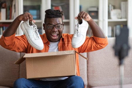 Téléchargez les photos : Émotionnel heureux cool jeune afro-américain gars avec dreadlocks blogueur de mode montrant disciples chaussures neuves blanches baskets élégantes, assis sur le canapé à la maison, streaming via smartphone - en image libre de droit
