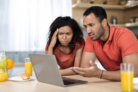 Photo for Sad unhappy black female and male in red t-shirts look at computer, have problems with gadget, service and bills, find mistake in kitchen interior. Bad news, stress, debts and bookkeeping at home - Royalty Free Image