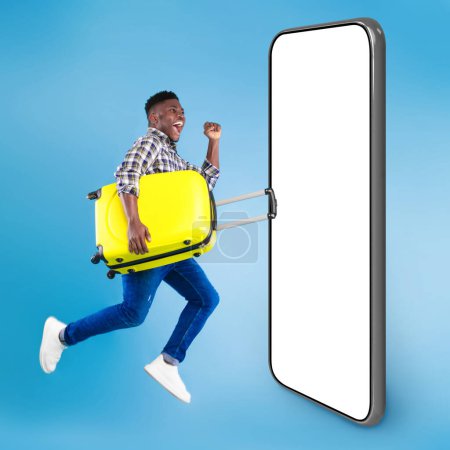 Téléchargez les photos : Jeune homme noir excité sautant avec une valise lumineuse par un grand smartphone avec écran blanc, faisant un geste OUI, remportant un voyage chaud en hiver sur fond bleu studio, une maquette, un collage - en image libre de droit