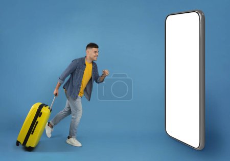 Photo for Full length portrait of cheerful male tourist with luggage running towards big phone with white screen over blue studio background. Young male passanger with baggage being making online reservation - Royalty Free Image
