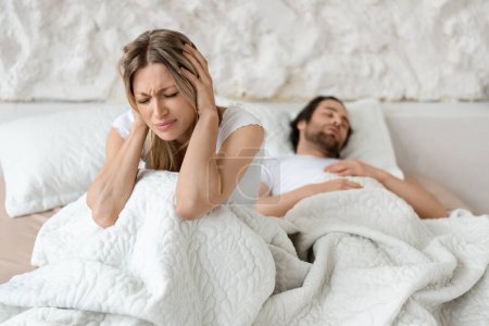 Foto de Mujer enojada infeliz sentada en la cama y cubriéndose las orejas con las manos, sufriendo de ronquidos de su marido, interior del dormitorio, espacio libre - Imagen libre de derechos