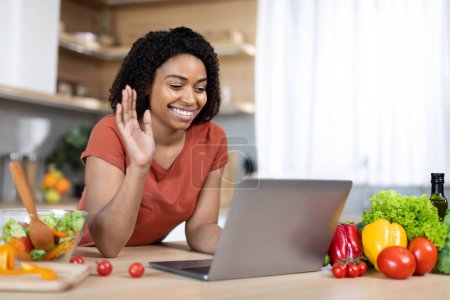 Photo for Smiling young black lady in red t-shirt preparing lunch, waving hand at computer webcam, shooting food blog in kitchen interior. Video call, meeting remotely, technology for communication at home - Royalty Free Image