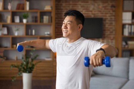 Foto de Estilo de vida deportivo. Hombre asiático maduro entrenando con pesas en casa, haciendo ejercicio en el interior de la sala de estar, disfrutando del entrenamiento doméstico. Feliz coreano masculino fortalecimiento de los músculos brazos - Imagen libre de derechos