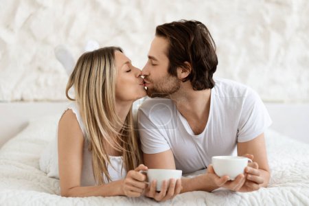 Foto de Amando pareja caucásica besándose mientras toman café en la cama del hotel, sosteniendo tazas, desayunando y disfrutando pasar tiempo juntos - Imagen libre de derechos