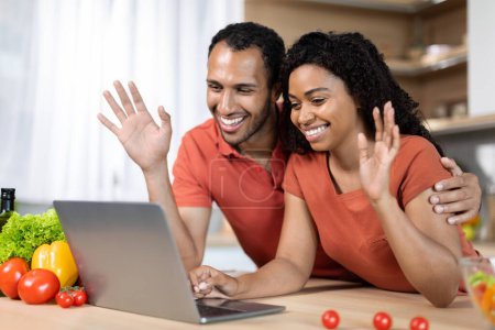 Photo for Cheerful young black guy and lady in red t-shirts have video call, waving hands at computer webcam in kitchen interior. Say hi, hello, greeting gesture during meeting remotely at home, covid-19 virus - Royalty Free Image