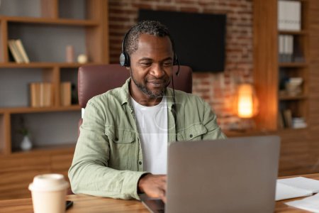 Photo for Glad mature black male in headset typing on computer at workplace with coffee takeaway, study and watch video in home office interior. Work and business with modern technology, meeting and video call - Royalty Free Image