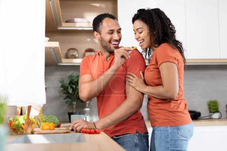 Photo for Cheerful millennial black male in red t-shirt cuts salad, feeds his wife in kitchen interior, copy space. Cooking meal together, relationships, romance and love, health care and homemade food at home - Royalty Free Image