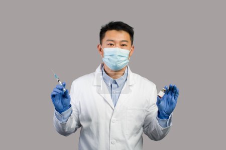 Photo for Serious mature chinese man therapist in white coat, protective mask shows vaccine and syringe, isolated on gray background. Vaccination immunization for health care, medical treatment due covid-19 - Royalty Free Image