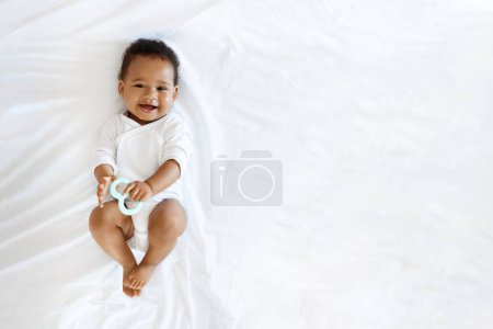 Photo for Childcare Concept. Portrait Of Adorable Little Black Baby Lying On Bed With Teether In Hand, Top View Shot Of Cute African American Infant Boy Or Girl Wearing Bodysuit Smiling At Camera, Copy Space - Royalty Free Image