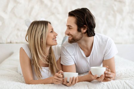 Photo for Morning drink in bed. Happy loving wife and husband lying on white bed with cups of hot coffee and looking at each other in bedroom interior, empty space - Royalty Free Image