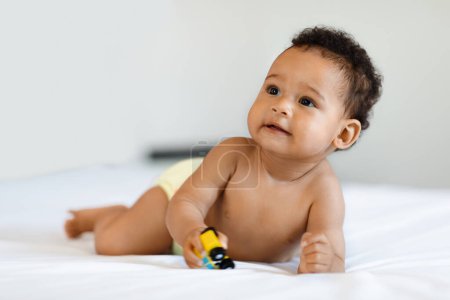 Foto de Adorable bebé negro que usa pañales jugando con el coche de juguete en la cama, lindo bebé afroamericano pequeño acostado sobre su barriga y mirando hacia otro lado, niño pequeño que se relaja en el dormitorio en casa, espacio de copia - Imagen libre de derechos