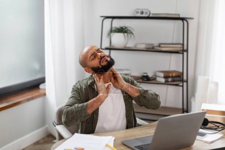 Photo for Upset latin male freelancer sitting at workplace at home, using laptop and rubbing his neck with both hands, suffering from pain while working online at home office - Royalty Free Image