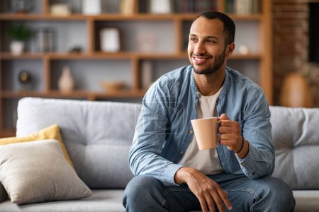 Téléchargez les photos : Portrait d'un bel homme afro-américain reposant sur un canapé à la maison avec une tasse de café, jeune homme noir assis sur un canapé dans le salon intérieur, regardant ailleurs et profitant d'une boisson chaude, copier l'espace - en image libre de droit