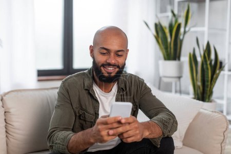 Foto de Hombre latino guapo usando teléfono inteligente mientras descansa en el sofá en casa, navegando por Internet o mensajería con amigos, disfrutando de las tecnologías modernas - Imagen libre de derechos