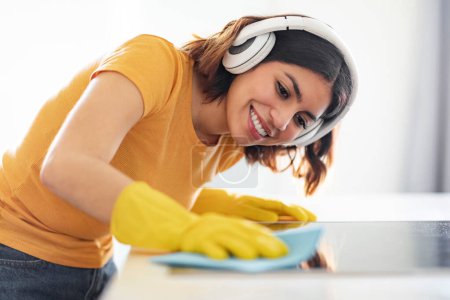 Foto de Joven árabe alegre escuchando música mientras limpia el mostrador de la cocina, sonriendo a la mujer de Oriente Medio usando auriculares inalámbricos y guantes de goma ordenando la superficie de la mesa con trapo, primer plano - Imagen libre de derechos