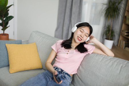 Photo for Pretty asian lady listening to music in modern wireless headphones, enjoying favorite songs with closed eyes, resting at home. Domestic entertainments, cool hobbies concept - Royalty Free Image