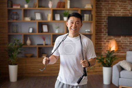 Photo for Excited asian middle aged man standing with skipping rope hang around his neck, ready to training at home in living room interior. Cardio active workout and healthy lifestyle - Royalty Free Image